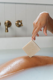 A person's hand holding a bar of soap, with the bar partially submerged in water, set against a bathroom backdrop with a bathtub and faucet.
