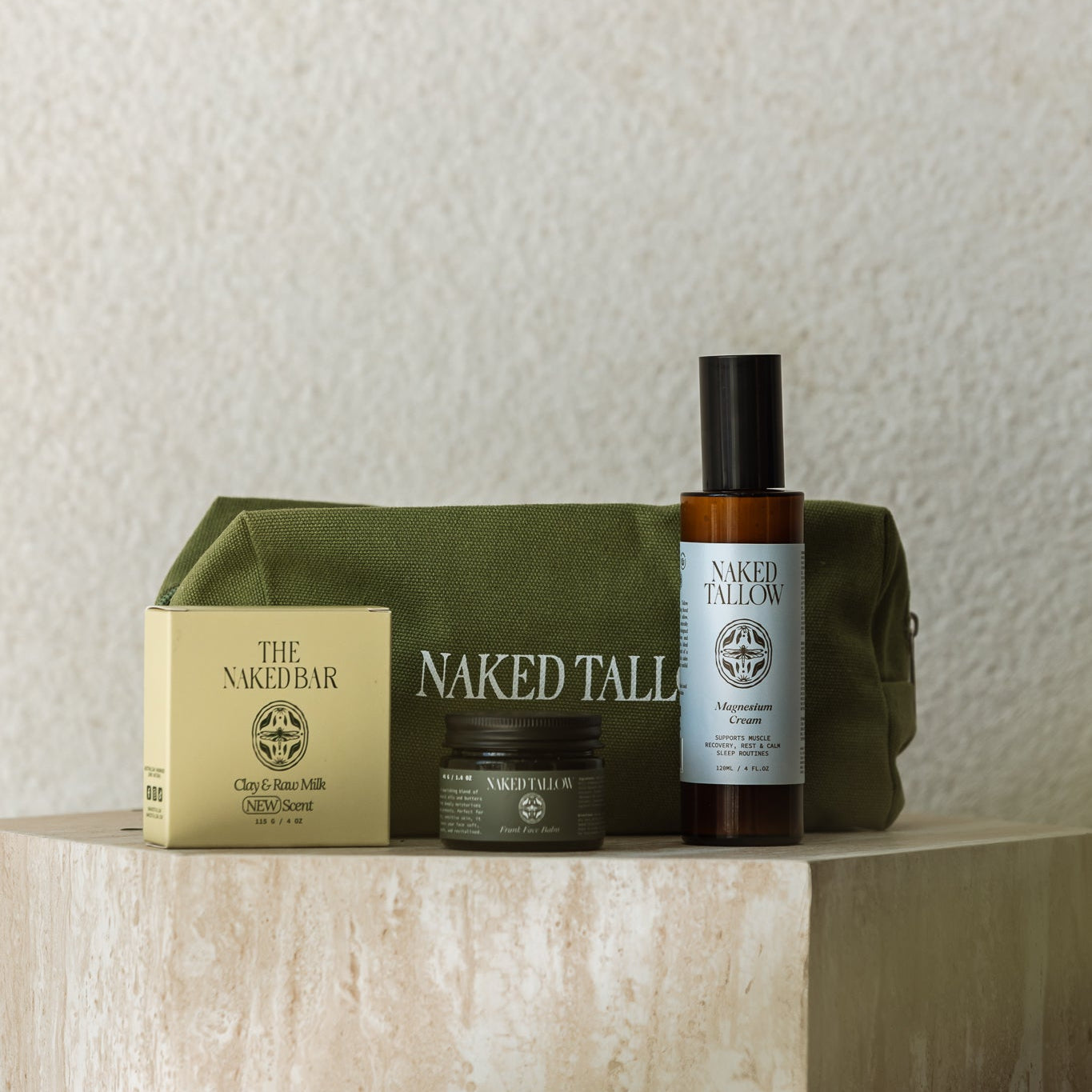 A curated product set from Naked Tallies and The Naked Bar arranged on a stone pedestal against a textured beige wall. The set includes an olive green zippered pouch, a gold-toned box, a dark amber bottle with a black cap, and a small jar.