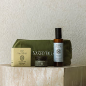 A curated product set from Naked Tallies and The Naked Bar arranged on a stone pedestal against a textured beige wall. The set includes an olive green zippered pouch, a gold-toned box, a dark amber bottle with a black cap, and a small jar.