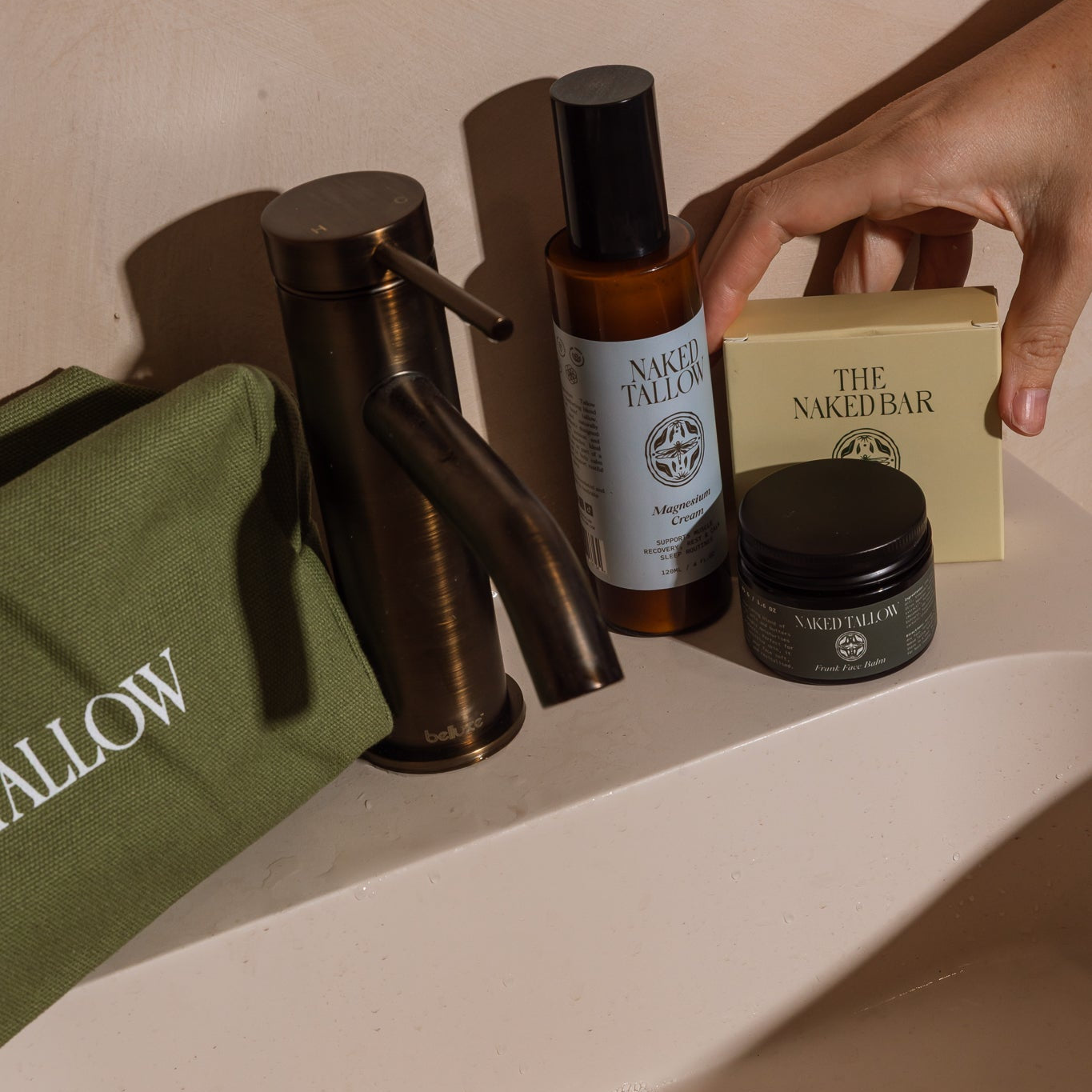 A hand reaches for a cream-colored bar of soap from The Naked Bar next to a bronze faucet, a green toiletry pouch, and an amber bottle on a bathroom ledge.