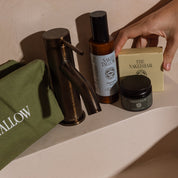 A hand reaches for a cream-colored bar of soap from The Naked Bar next to a bronze faucet, a green toiletry pouch, and an amber bottle on a bathroom ledge.
