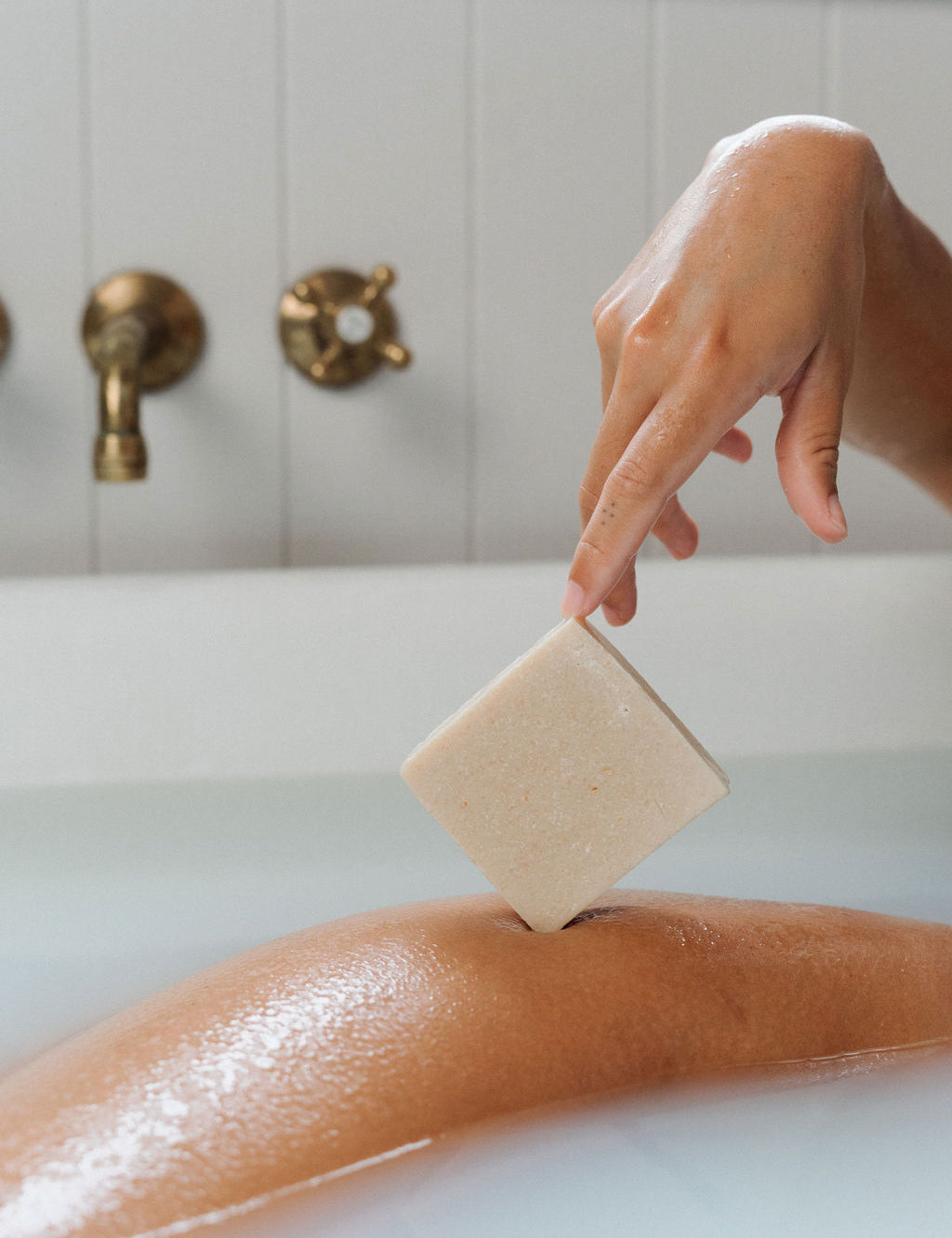 A person's hand holding a bar of soap, with the bar partially submerged in water, set against a bathroom backdrop with a bathtub and faucet.