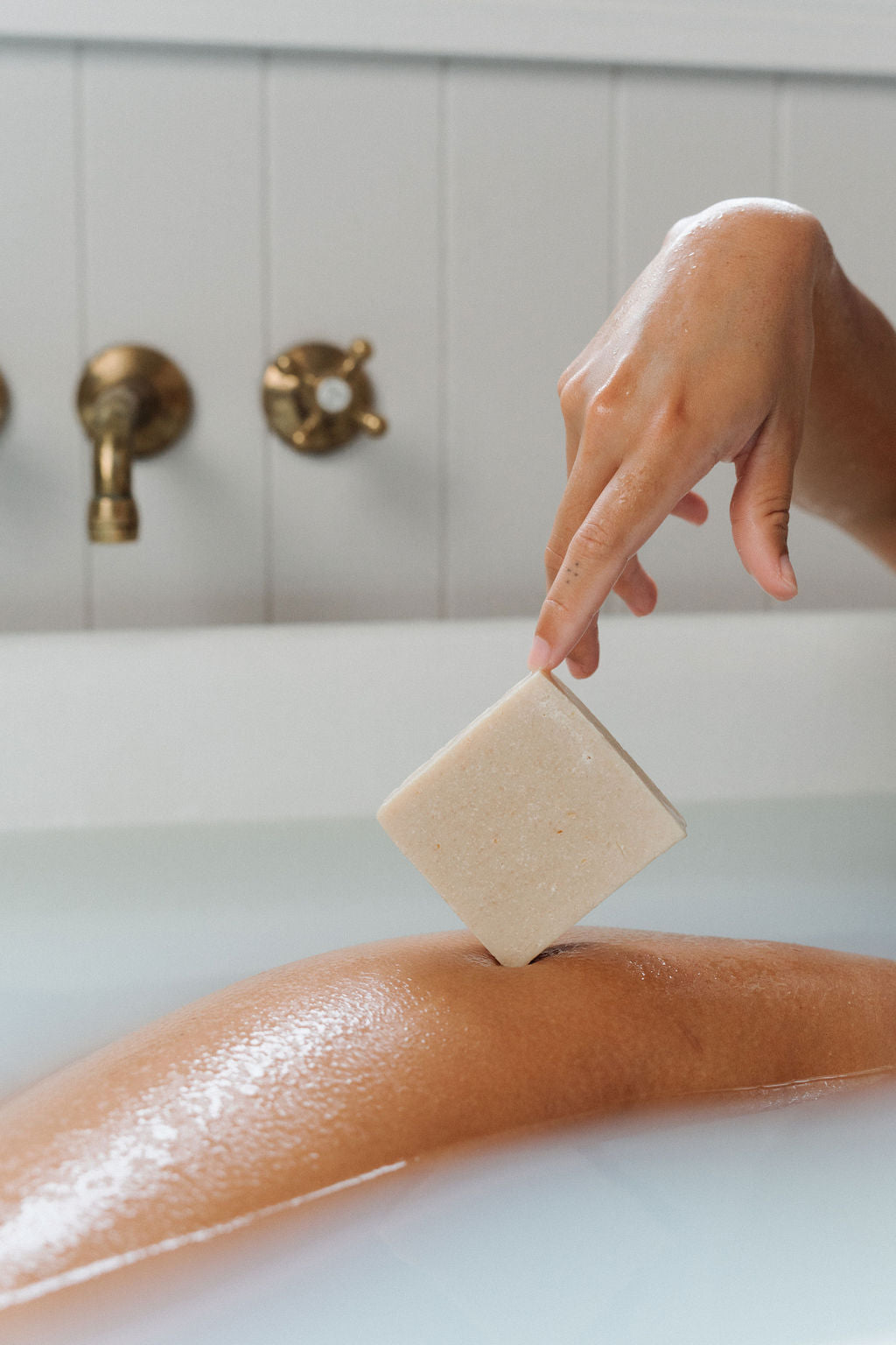 A person&#39;s hand holding a bar of soap, with the bar partially submerged in water, set against a bathroom backdrop with a bathtub and faucet.
