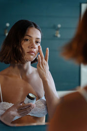 Woman applying cream to her face in front of a mirror - beef tallow for skin hydrating face balm by naked tallow.