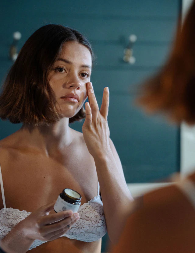 Woman applying cream to her face in front of a mirror - beef tallow for skin hydrating face balm by naked tallow.