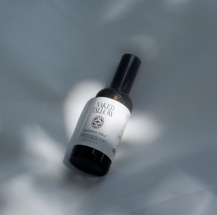 A bottle of Hydrating Mist by Naked Tallow, placed on a surface with a shadow falling over it.
