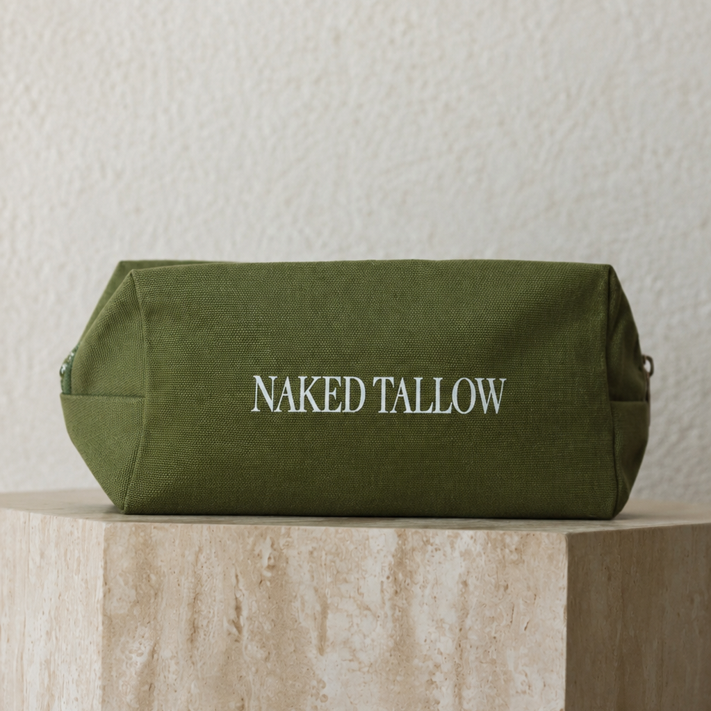 An olive green canvas cosmetic pouch with 'NAKED TALLOW' printed in white serif letters, displayed on a textured beige stone pedestal against a matching textured wall.