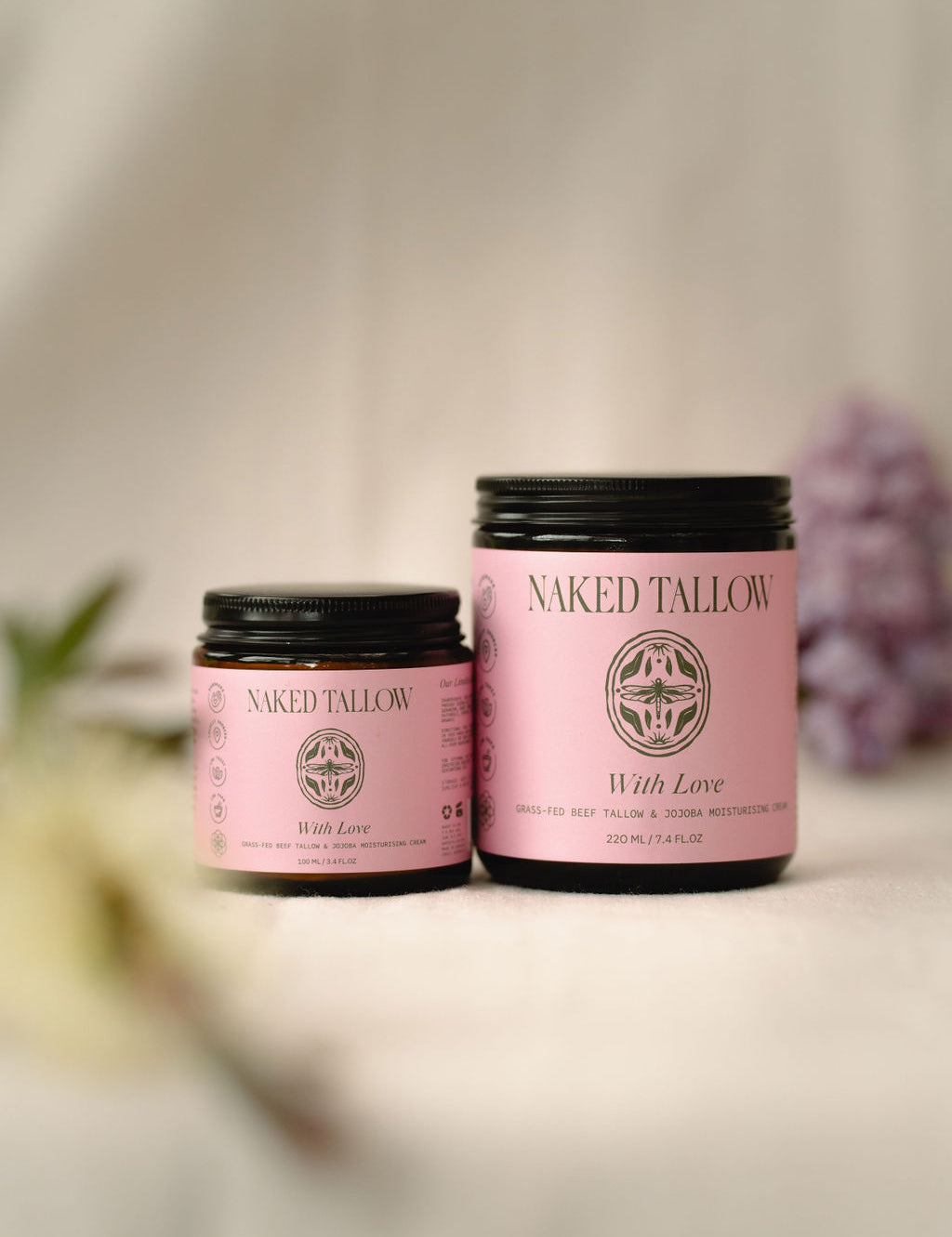 Two jars of Naked Tallow 'With Love' Tallow Cream, one small and one large, with a pink label and floral imagery, displayed on a soft, light background with a sprig of dried flowers to the side.
