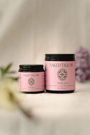 Two jars of Naked Tallow 'With Love' Tallow Cream, one small and one large, with a pink label and floral imagery, displayed on a soft, light background with a sprig of dried flowers to the side.